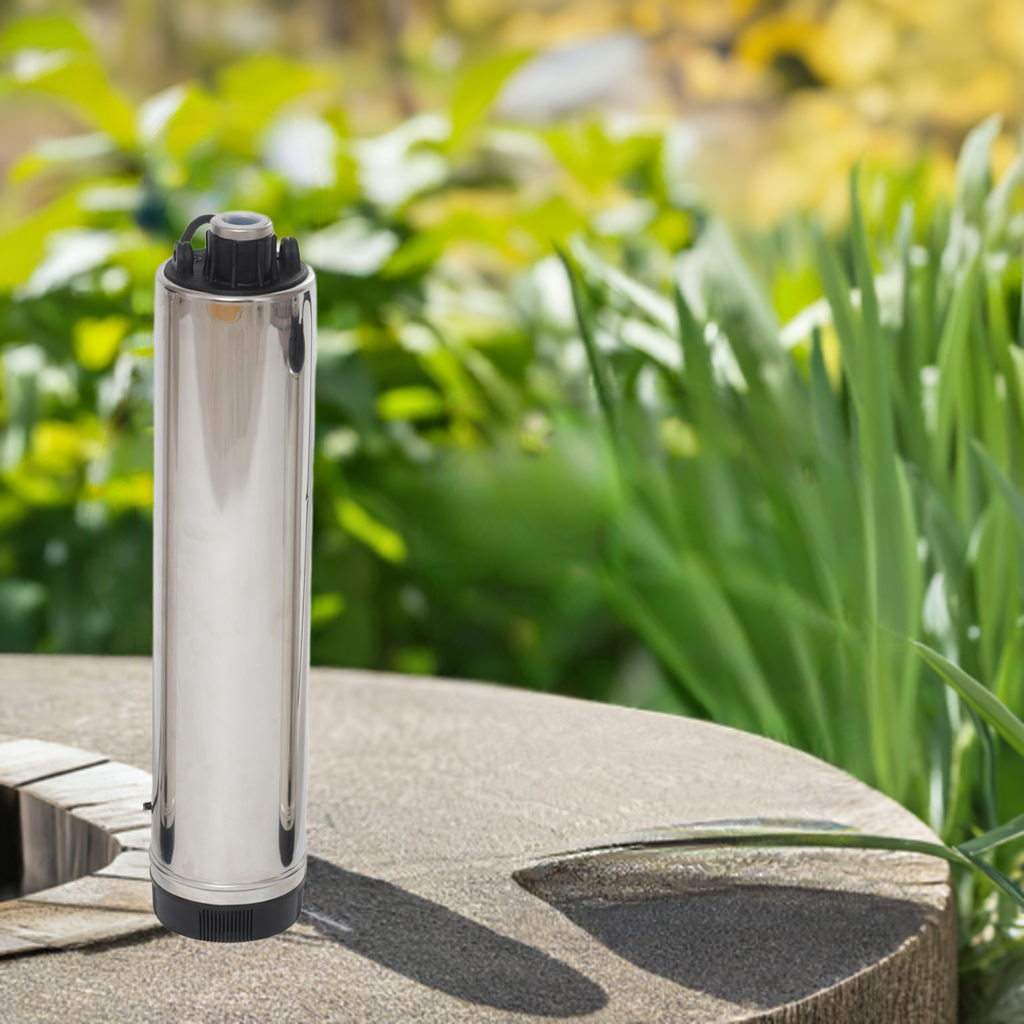 A vertical, silver-colored submersible pump is shown, made of stainless steel, placed on a stone surface outdoors with greenery in the background.